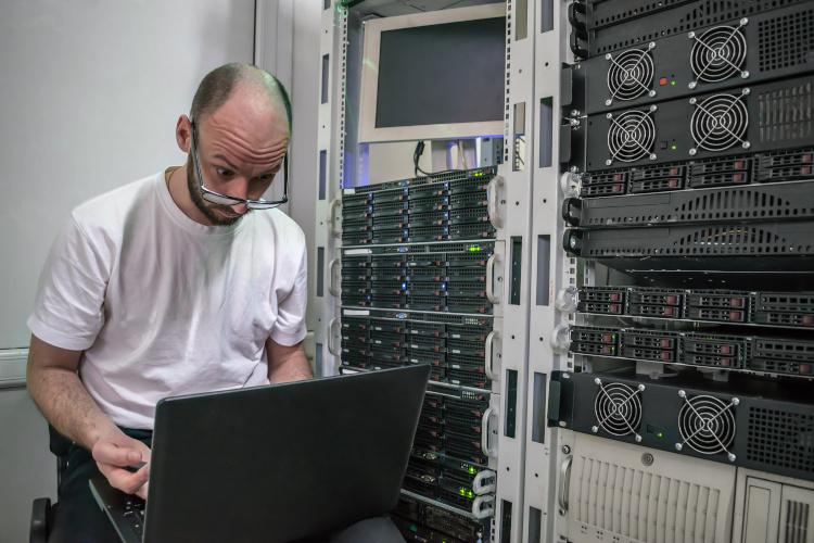 Surprised sysadmin looking at their laptop while sitting in front of a rack of servers